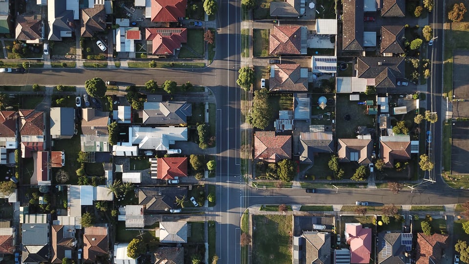 Aerial view of a suburban neighborhood, various houses, green lawns, intersecting roads, bathed in soft sunlight.