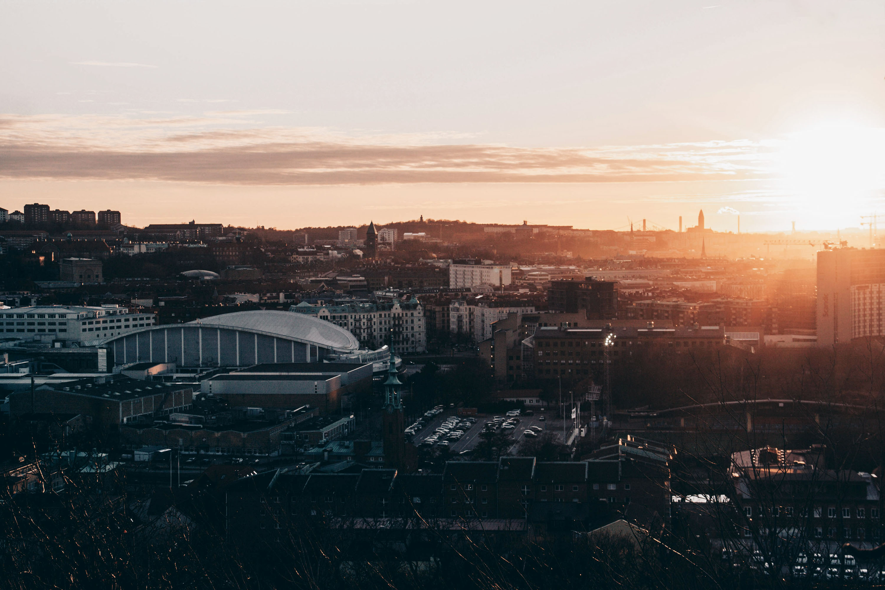 A cityscape at sunset with a prominent stadium in the foreground, buildings bathed in warm light, and hills in the distance.