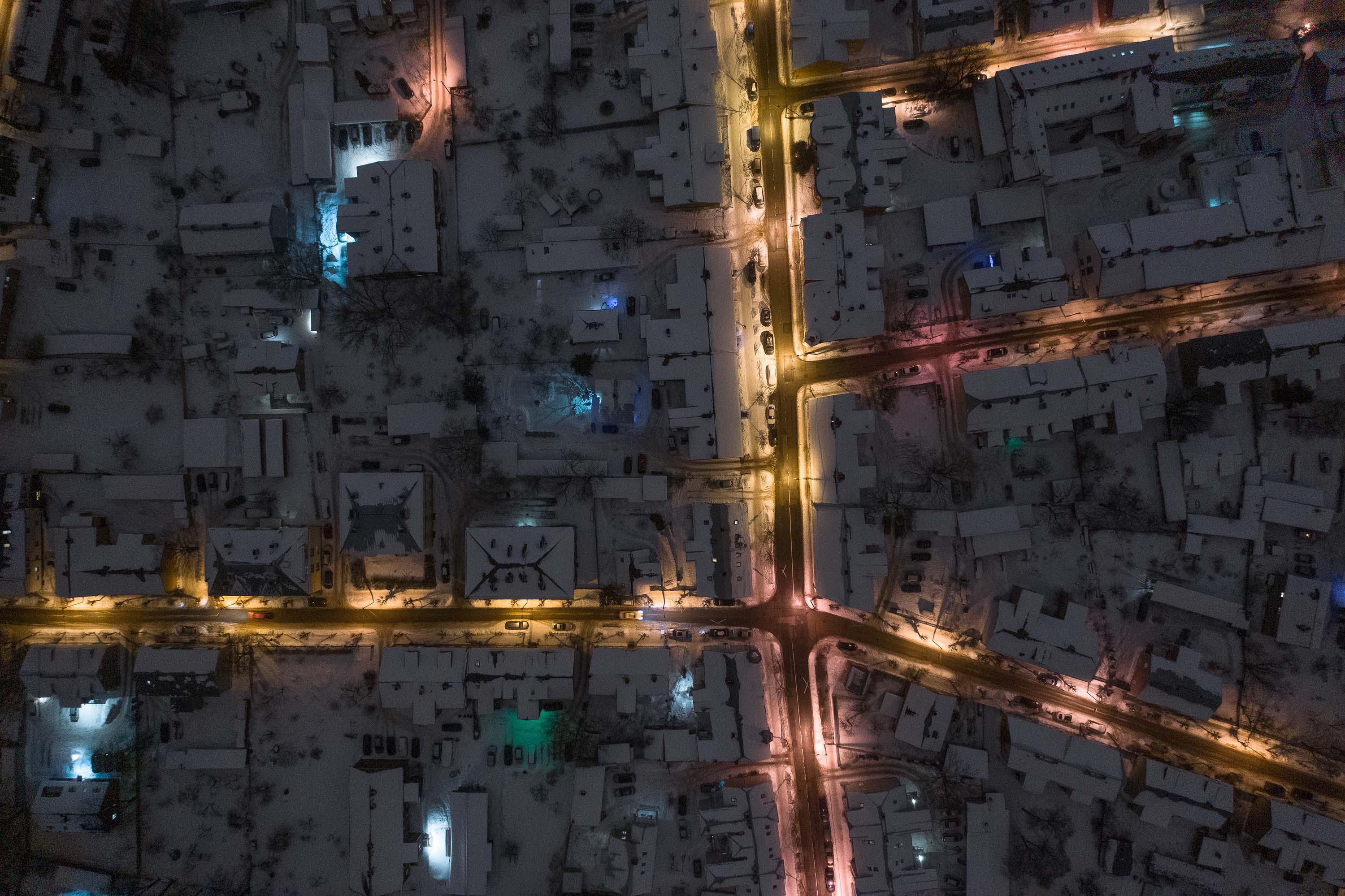 Aerial view of a city at night, with illuminated roads forming a bright network amidst snow-covered, darkened buildings.