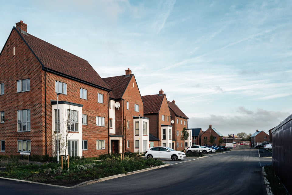 A peaceful neighborhood with modern brick houses, greenery, parked cars, under a partly cloudy sky.