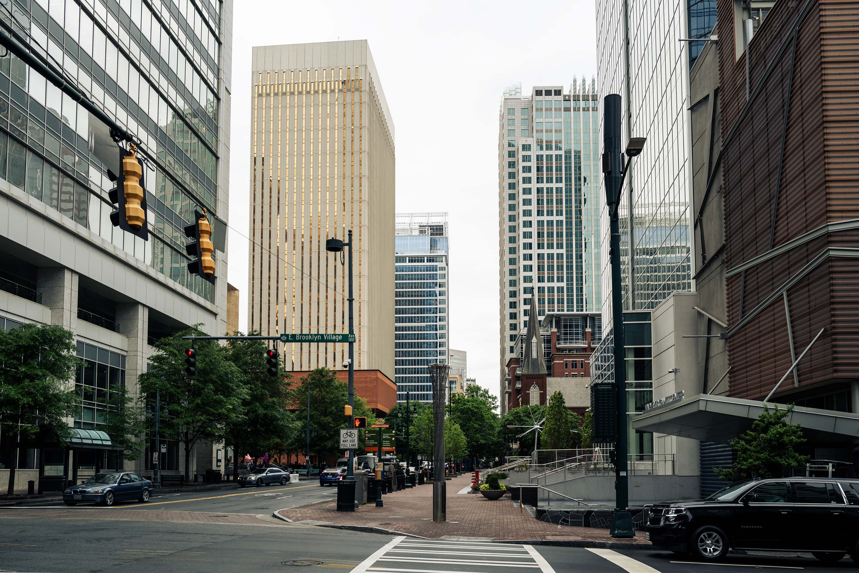 A city intersection with diverse tall buildings, traffic lights, a parked car, pedestrian pathways, and small greenery. Cloudy sky above.